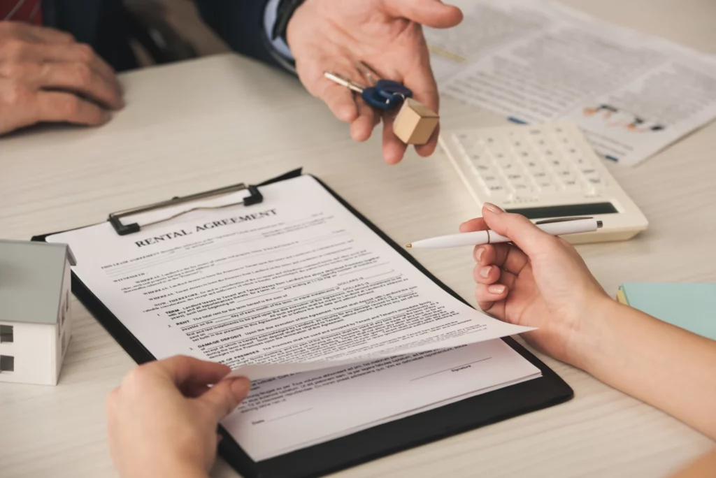 A rental agreement on a clipboard being reviewed, with one hand holding a pen and another offering keys. A small house model and calculator are nearby.