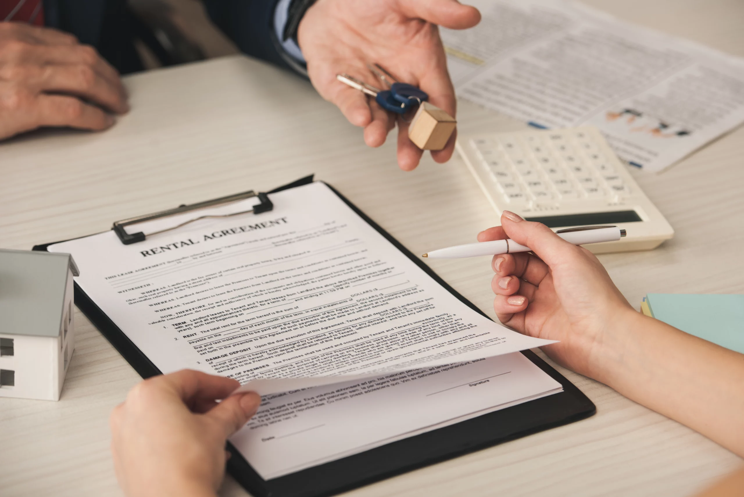 A rental agreement on a clipboard being reviewed, with one hand holding a pen and another offering keys. A small house model and calculator are nearby.