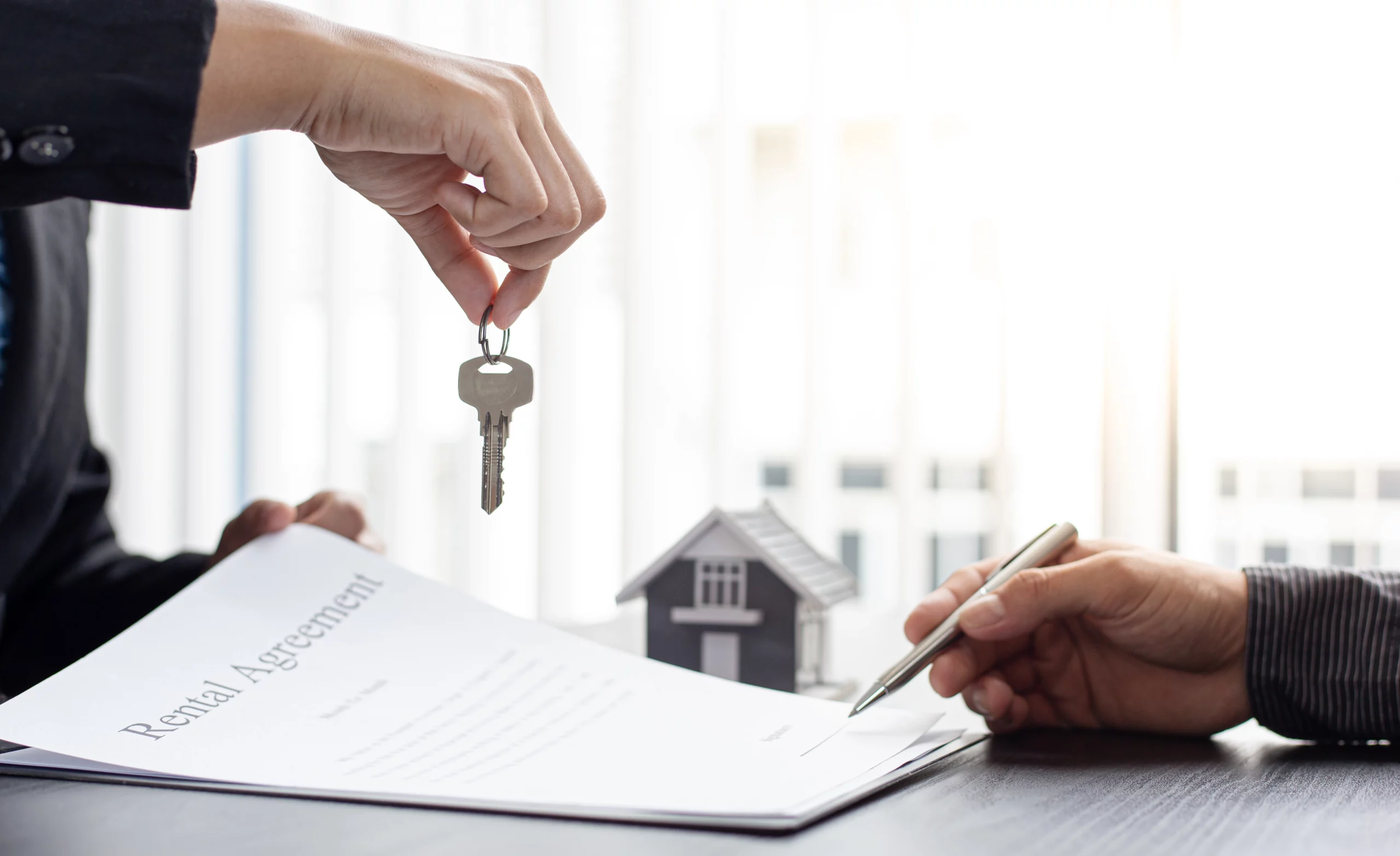 Hands exchanging house keys over a rental agreement as a person signs the lease, with a small house model nearby